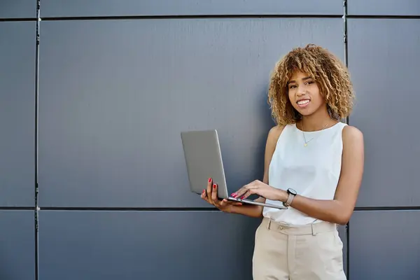 Feliz encaracolado Africano americano freelancer de pé com seu laptop perto de parede cinza do edifício de escritórios — Fotografia de Stock