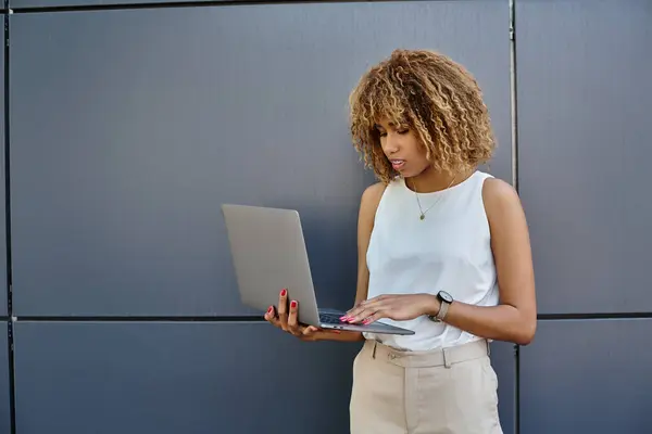 Ricci afroamericani donna d'affari con bretelle in piedi con il suo computer portatile vicino al muro grigio di costruzione — Foto stock