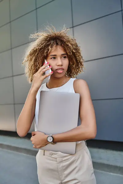 Moderna joven profesional, mujer afroamericana con portátil haciendo una llamada contra la pared gris - foto de stock