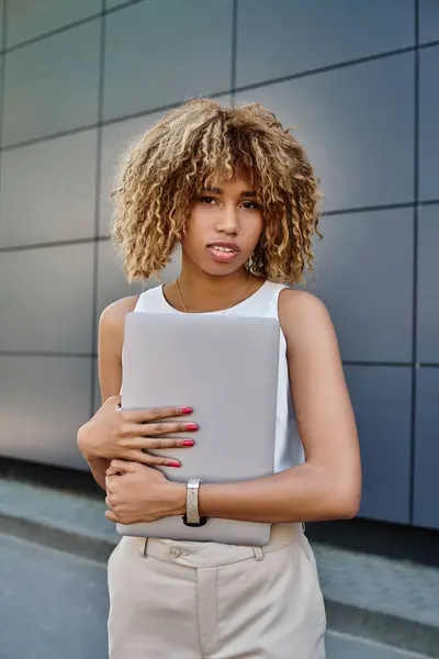 Ricci afroamericani donna d'affari con bretelle in piedi con il suo computer portatile vicino edificio per uffici — Foto stock