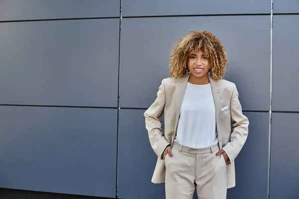 Jovem mulher de negócios negra feliz em terno de pé com as mãos nos bolsos contra edifício cinza — Fotografia de Stock