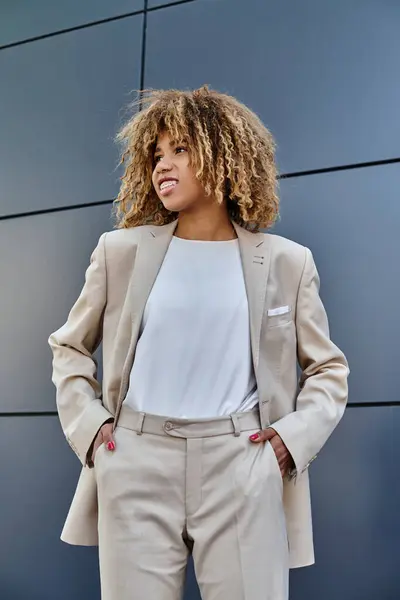 Heureuse femme d'affaires afro-américaine en costume debout avec les mains dans les poches contre le bâtiment gris — Photo de stock