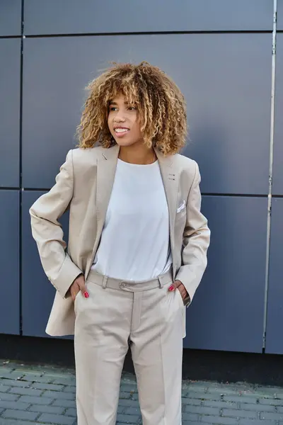 Fröhliche afrikanisch-amerikanische Geschäftsfrau im Anzug, die Hände in den Taschen vor dem grauen Gebäude — Stockfoto