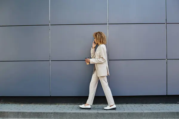 Femme d'affaires afro-américaine bouclée marchant avec du café et parlant par téléphone contre un mur gris — Photo de stock