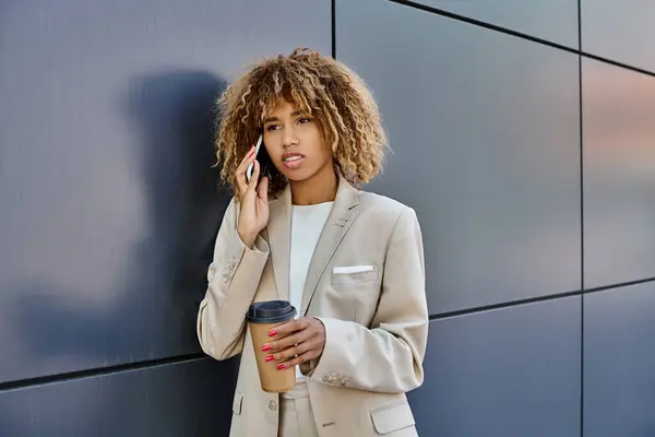 Curly african american businesswoman standing with coffee and talking by phone against grey wall — Stock Photo