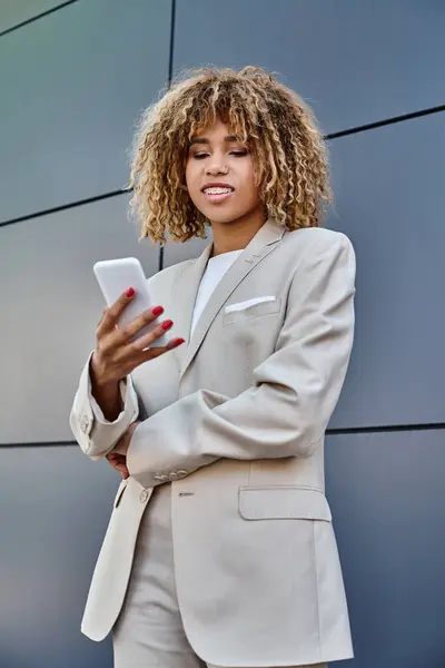 Позитивная и молодая африканская американская бизнесвумен с вьющимися волосами держит свой смартфон на открытом воздухе — стоковое фото