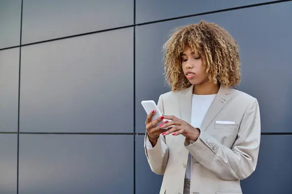 Молода афроамериканська бізнес-леді з кучерявим волоссям, що стоїть в костюмі і використовує свій смартфон — стокове фото