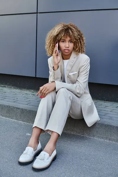 Young african american businesswoman in suit sitting on parapet and talking on her smartphone — Stock Photo