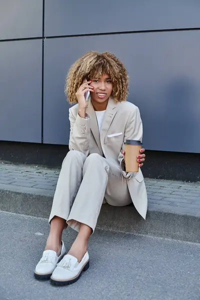 African american woman in suit sitting on parapet with coffee to go and talking on her smartphone — Stock Photo
