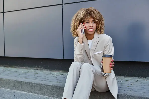 Young african american woman in suit sitting on parapet with coffee to go and talking on smartphone — Stock Photo