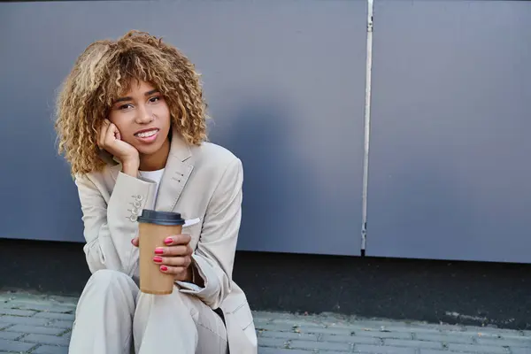 Felice donna afroamericana in giacca e cravatta seduta sul parapetto con caffè per andare a guardare la macchina fotografica — Foto stock
