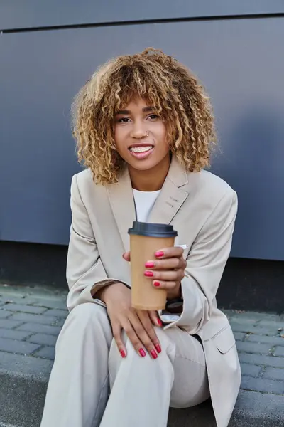 Sorridente donna afroamericana in completo seduta su parapetto con caffè per andare a guardare la macchina fotografica — Foto stock