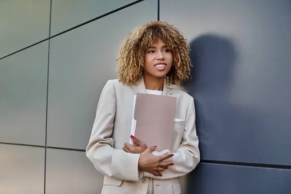 Cheerful young african american businesswoman in formal wear holding folder by grey office wall — Stock Photo