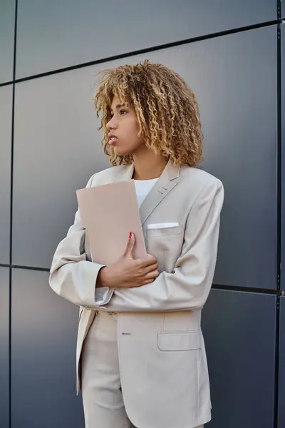 Curly and young african american businesswoman in formal wear holding folder by grey office wall — Stock Photo