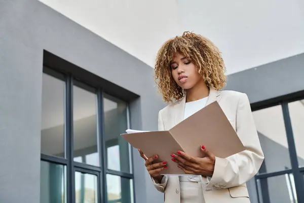 Enfocado africano americano rizado mujer de negocios revisar el papeleo en carpeta cerca de edificio de oficinas - foto de stock