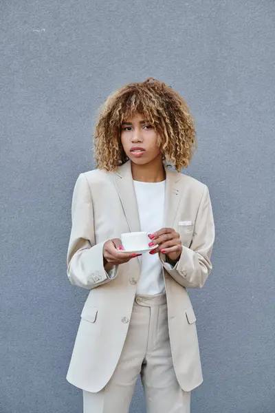 Curly african american businesswoman posing with cup of coffee near grey wall of office building — Stock Photo