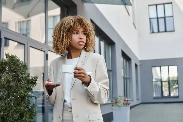 Encaracolado Africano americano empresária de pé com xícara de café perto da entrada do prédio de escritórios — Fotografia de Stock