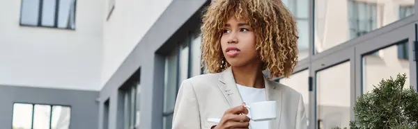 Mujer de negocios afroamericana rizada de pie con taza de café cerca del edificio de oficinas, pancarta - foto de stock