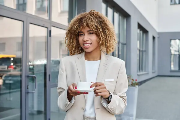 Femme d'affaires afro-américaine souriante dans la vingtaine, debout avec une tasse de café près d'un immeuble de bureaux — Photo de stock