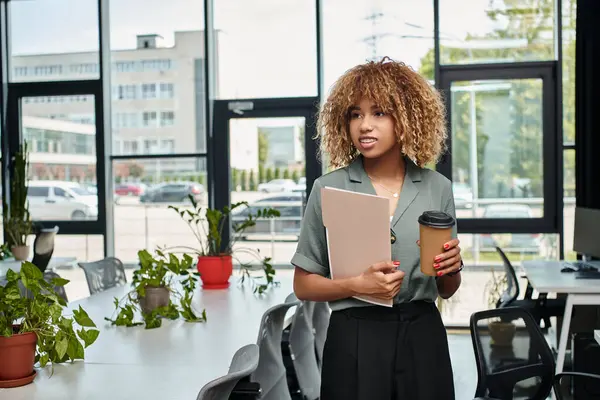 Curly african american businesswoman in her 20s, standing with coffee and folder in office — Stock Photo