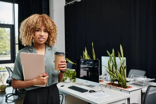 Femme d'affaires afro-américaine frisée dans la vingtaine, debout avec du café à emporter et un dossier au bureau — Photo de stock