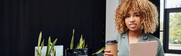 Curly african american businesswoman in her 20s, standing with coffee and folder in office, banner — Stock Photo
