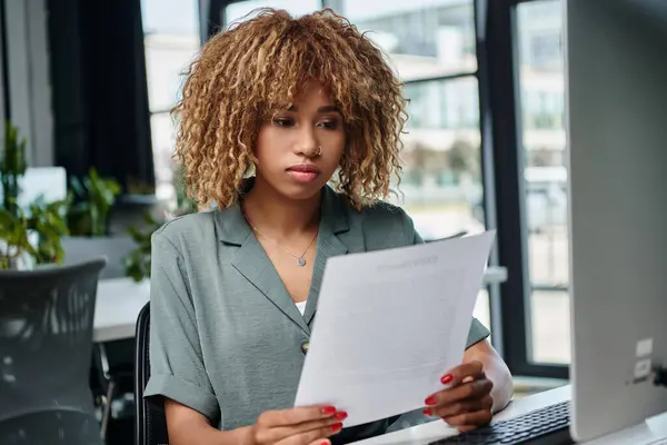 Curly young african american manager reviewing document in front of computer monitor in office — Stock Photo