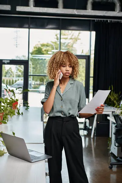 Багатозадачна афроамериканська бізнес-леді з документом, що приймає дзвінок біля ноутбука в сучасному офісі — стокове фото