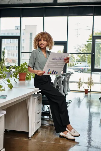 Joyeuse femme d'affaires afro-américaine évaluant les graphiques dans un espace de bureau dynamique, gestionnaire — Photo de stock