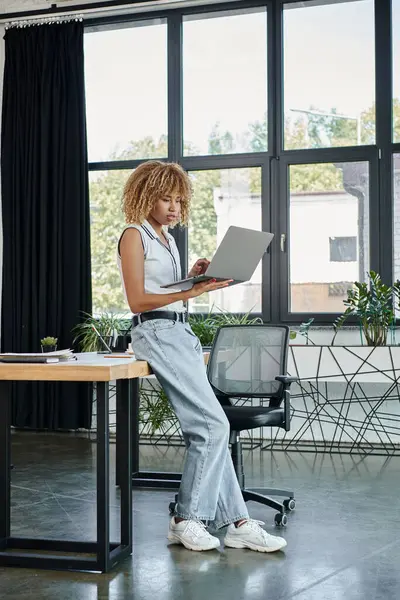 Mujer de negocios afroamericana rizada en traje casual trabajando en el ordenador portátil en una configuración de oficina moderna - foto de stock