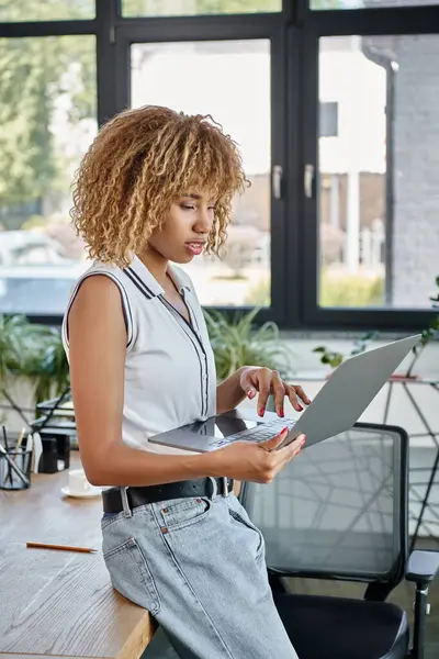Frisée afro-américaine femme d'affaires en tenue décontractée travaillant sur ordinateur portable dans un espace de travail moderne — Photo de stock