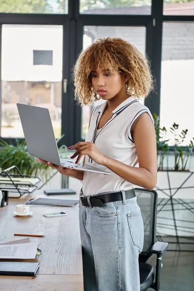 Mujer de negocios afroamericana rizada con atuendo casual trabajando en una computadora portátil en una oficina moderna - foto de stock