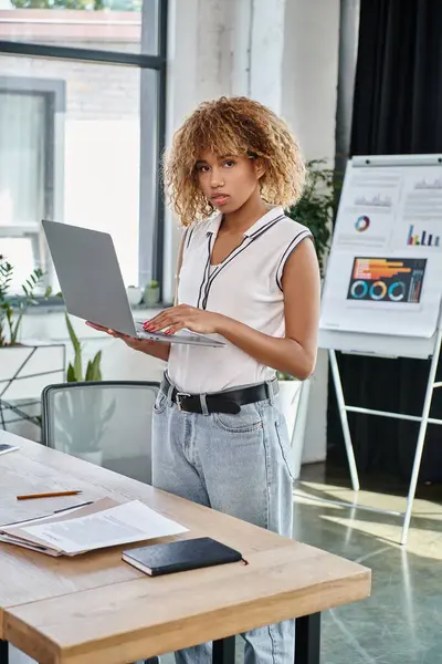 Jeune femme d'affaires afro-américaine frisée en tenue décontractée travaillant sur ordinateur portable dans un bureau moderne — Photo de stock
