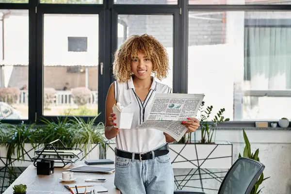 Happy african american woman holding asian takeaway meal in carton box and newspaper in office — Stock Photo