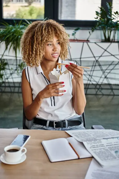 Happy curly african american woman holding chopstick with takeaway noodles in carton box at desk — Stock Photo