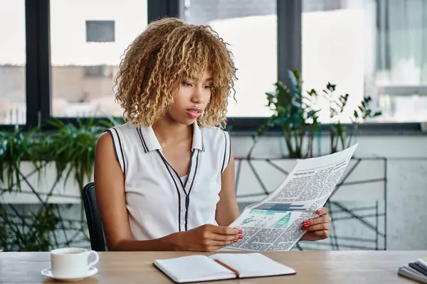 Mujer de negocios afroamericana rizada leyendo el periódico y sentada en el escritorio en la oficina, profesión - foto de stock