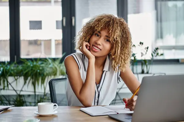 Feliz mujer de negocios afroamericana con frenos tomando notas al lado de la computadora portátil en la oficina moderna - foto de stock