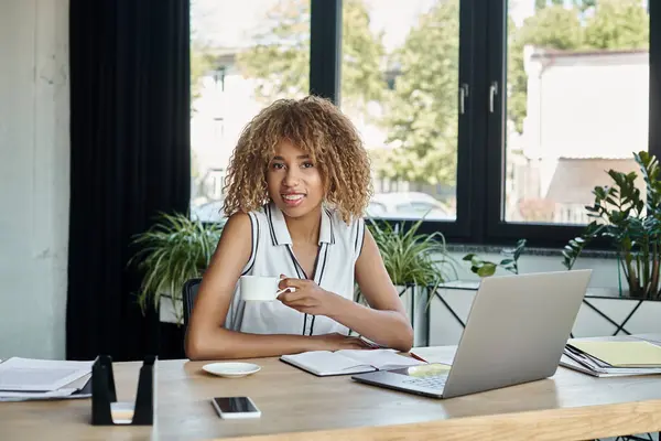 Feliz mujer de negocios afroamericana con frenos sosteniendo la taza de café al lado de la computadora portátil en la oficina - foto de stock
