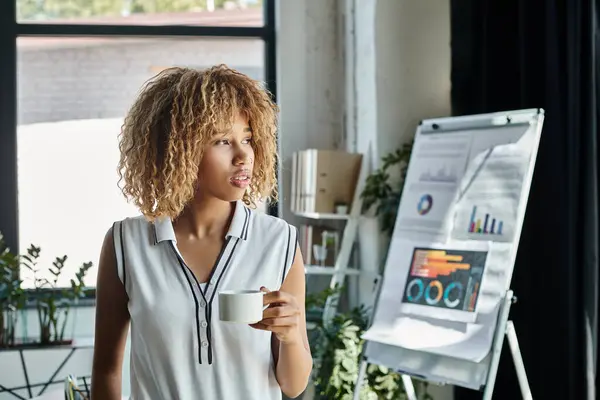 Curly african american businesswoman with braces holding cup of coffee beside charts and graphs — Stock Photo