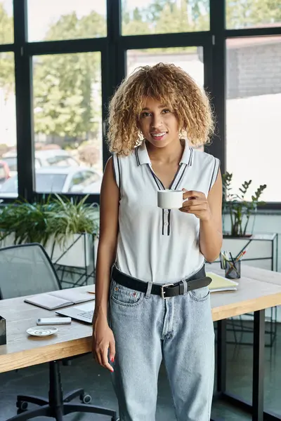 Feliz mulher de negócios afro-americana com aparelho segurando xícara de café ao lado da mesa de trabalho — Fotografia de Stock