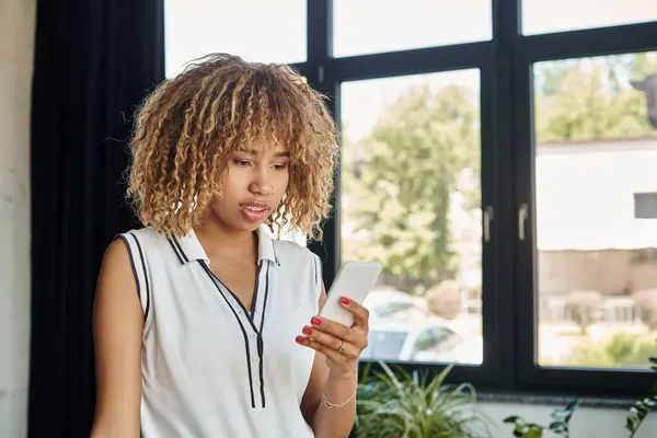 Mujer de negocios afroamericana rizada y joven con teléfono inteligente permaneciendo conectado en la oficina iluminada por el sol - foto de stock