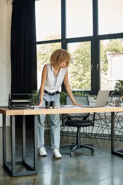 Focada jovem afro-americana de pé em sua mesa e olhando para laptop durante o dia de trabalho — Fotografia de Stock