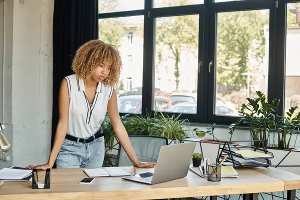 Concentrée femme afro-américaine bouclée debout à son bureau et regardant ordinateur portable pendant la journée de travail — Photo de stock