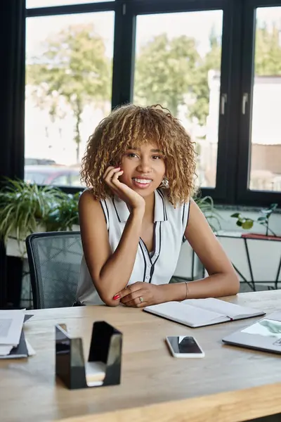 Feliz joven mujer de negocios afroamericana sonriendo cerca de la computadora portátil y teléfono inteligente en el escritorio de trabajo - foto de stock