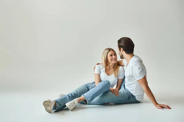 Un uomo e una donna, che incarnano amore e vicinanza, si siedono a terra in studio grigio — Foto stock