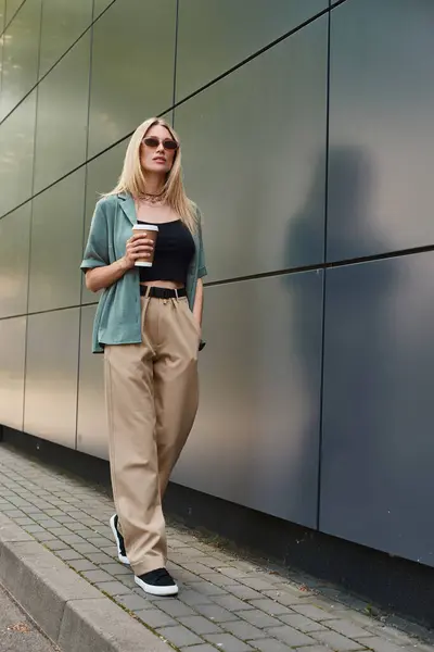 Una mujer vestida con ropa casual de pie junto a una pared de ladrillo, sosteniendo una taza de café y tomando un momento para saborear la bebida. - foto de stock