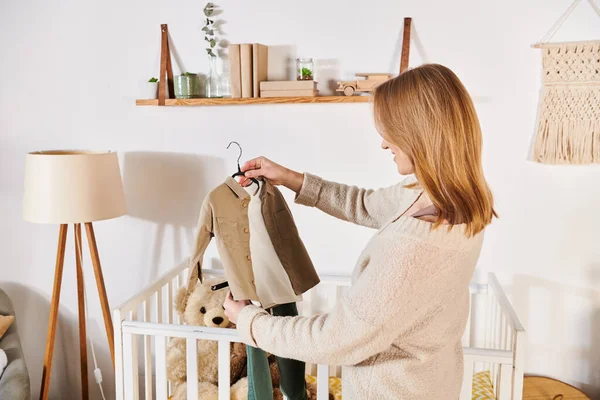 Jovem futura mãe segurando roupas de bebê perto berço com brinquedos macios no quarto do berçário, expectativa de nascimento — Fotografia de Stock