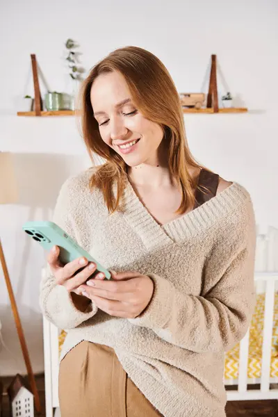 Mensajería de mujer embarazada sonriente en el teléfono móvil en la habitación de la guardería en casa, expectativa de maternidad - foto de stock