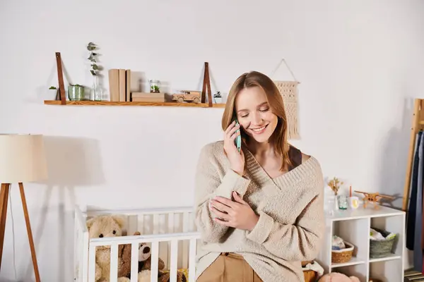 Jovem alegre falando no telefone celular perto do berço com brinquedos suaves no quarto de crianças, futura mãe — Fotografia de Stock