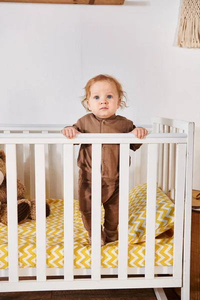 Adorabile bambino bambino in pagliaccetto in piedi in culla nella stanza dei bambini a casa, infanzia felice — Foto stock